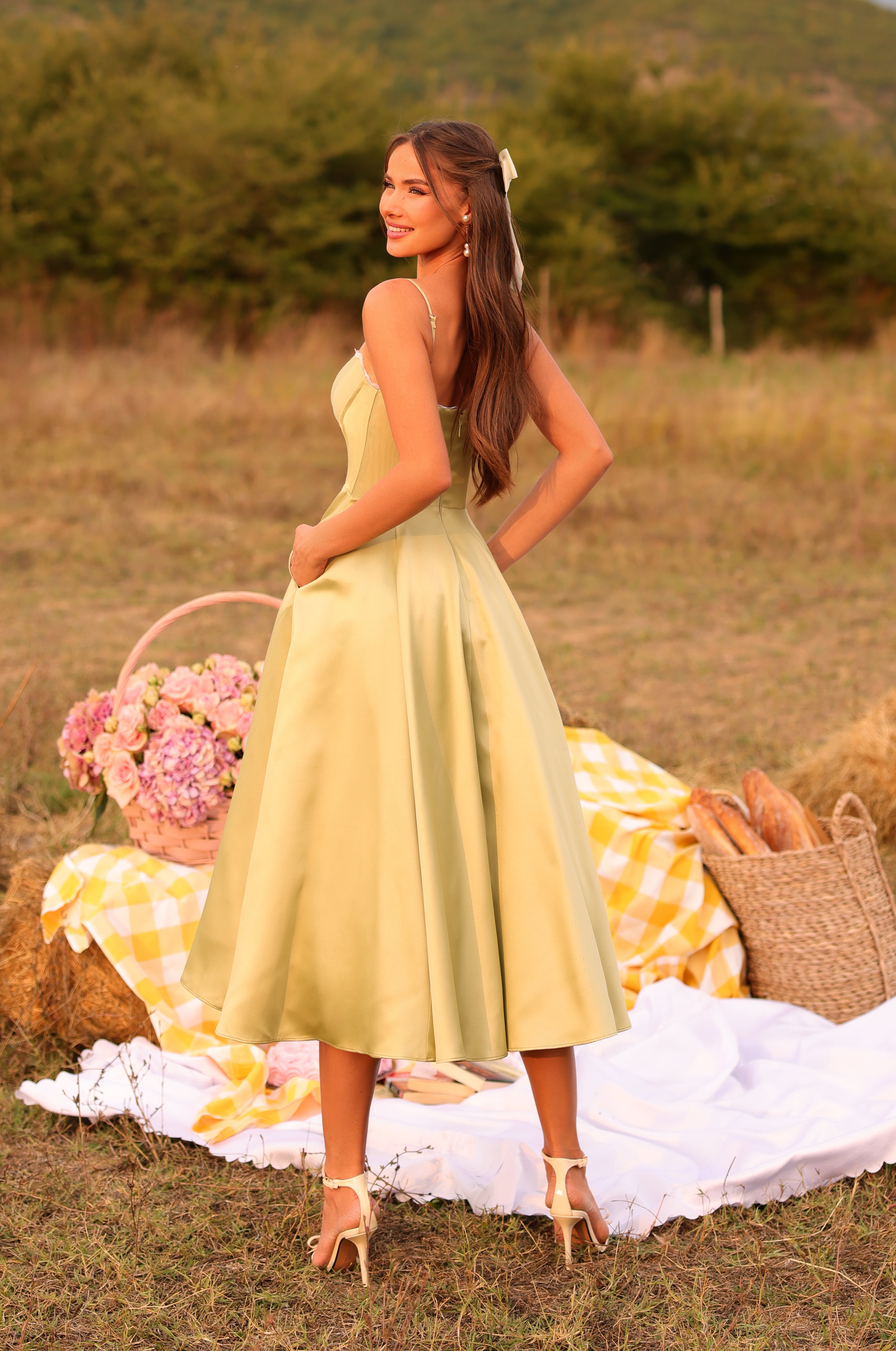 A woman in the Olive Green Satin Midi Wedding Guest Dress: Olivetta stands on a picnic setup outdoors, with a basket of flowers and bread on a blanket and hay bale in the background.